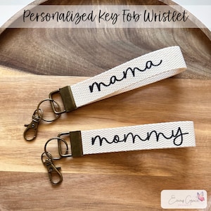 May include: Two white canvas wristlets with keychains. The wristlets are personalized with the words "mama" and "mommy" in black cursive lettering.