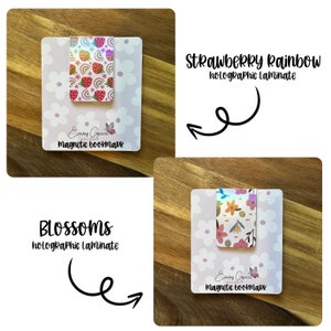 Puede incluir: Dos marcadores magn&eacute;ticos con acabado laminado hologr&aacute;fico. El primer marcador presenta un dise&ntilde;o de fresa y arco&iacute;ris con el texto "Strawberry Rainbow holographic Laminate". El segundo marcador presenta un dise&ntilde;o floral con el texto "Blossoms holographic Laminate". Ambos marcadores tienen el texto "Emmy Grace Designs Magnetic Bookmark" en la parte inferior.