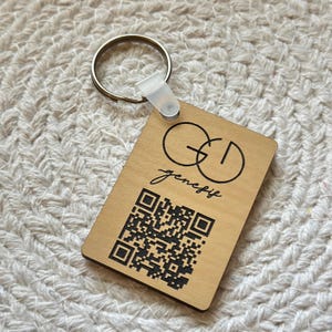 May include: A rectangular wooden keychain with a gold-colored ring and a clear plastic tag. The keychain features the text "GD genesif" and a black QR code. The wood has a light brown color.