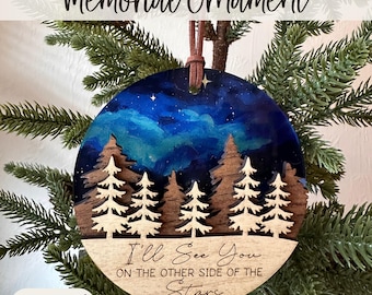 May include: A round wooden memorial ornament with a painted night sky scene and laser-cut trees. The text "I'll See You on the Other Side of the Stars" is inscribed on the bottom. A brown leather cord is attached for hanging.