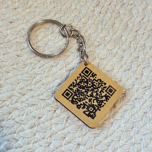 May include: A wooden keychain with a silver metal ring and chain. The keychain has a diamond shape with a QR code printed on it. The background is a light beige woven surface.