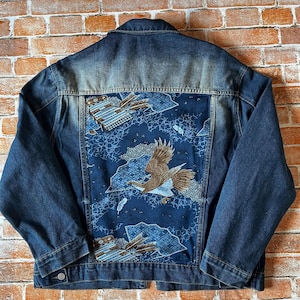 Puede incluir: Una chaqueta de mezclilla azul oscuro con un gran panel trasero rectangular con un diseño estampado. El panel muestra un águila marrón en vuelo, flechas estilizadas y patrones geométricos en tonos azul, blanco y marrón sobre un fondo azul.