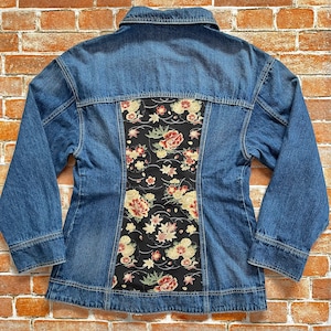 Op de afbeelding: Een blauwe spijkerjas met een bloemenmotief op de rug. Het bloemmotief heeft rode, gele en witte bloemen op een zwarte achtergrond. De jas heeft lange mouwen en een knoopsluiting.