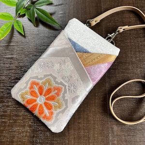 Könnte beinhalten: Eine weiße und silberne Stoff-Umhängetasche mit Blumenmuster. Die Tasche hat einen langen Riemen und einen Klappenverschluss. Die Tasche ist aus einem Patchwork aus verschiedenen Stoffen, darunter Blau, Rosa und Gold.