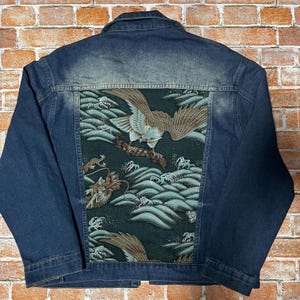 Könnte beinhalten: Eine dunkelblaue Jeansjacke mit einem großen rechteckigen Rückenteil aus dunkelgrünem Stoff. Der Einsatz zeigt einen detaillierten Grafikdruck eines braunen Adlers im Flug über stilisierten hellgrünen Meereswellen und einem Drachen.