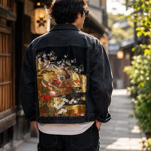 May include: A black denim jacket featuring a large, rectangular back panel with a detailed gold-toned illustration of birds, floral blossoms, and traditional decorative elements. The jacket has a classic collar, long sleeves, and a button-up front.
