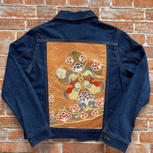 Peut inclure: Une veste en jean bleu foncé avec un grand panneau rectangulaire au dos. Le panneau est en tissu brun doré avec un motif brodé de fleurs stylisées et de vagues en fils de couleur blanche, rouge, jaune et violet foncé.
