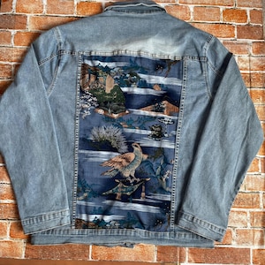 Könnte beinhalten: Eine hellblaue Jeansjacke mit einem großen rechteckigen Rückenteil mit aufgedruckter Grafik. Die Grafik zeigt einen Raubvogel, der auf einer Struktur sitzt, umgeben von stilisierten Landschaftselementen, Bäumen und einer Teekanne in Blau- und Grüntönen.
