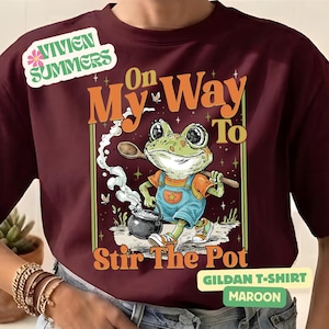 Puede incluir: Camiseta Gildan granate con un dibujo de una rana de dibujos animados removiendo una olla. El texto dice "On My Way To Stir The Pot". La rana lleva un peto azul y zapatos naranjas.