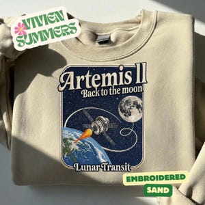 Puede incluir: Sudadera de color arena con un diseño gráfico. El diseño presenta el texto "Artemis II Back to the moon!" y una ilustración de temática espacial. El diseño incluye un satélite, la Tierra, la luna y las palabras "Lunar Transit". La sudadera también tiene el texto "Embroidered Sand".