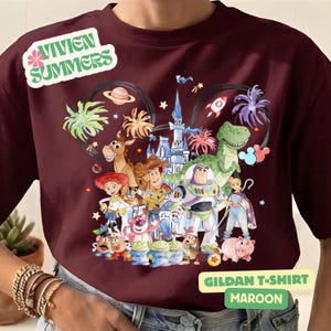 Puede incluir: Camiseta Gildan granate con un gr&aacute;fico colorido de los personajes de Toy Story. El dise&ntilde;o incluye a Woody, Buzz Lightyear y otros personajes, con fuegos artificiales y un castillo. La camiseta tiene el texto "Vivien Summers".