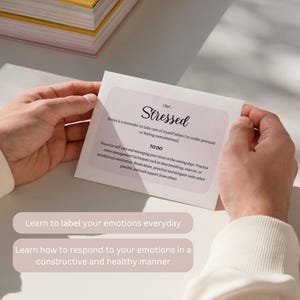 May include: A person holding a white card with the word "Stressed" in elegant script. The card includes a list of "To Do" items for managing stress, with text that reads "Learn to label your emotions everyday" and "Learn how to respond to your emotions."