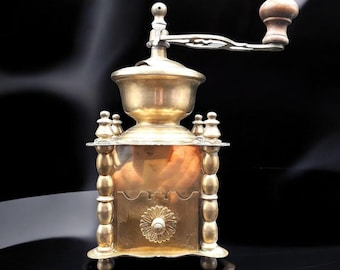 May include: Antique brass coffee grinder with a wooden handle and ornate details. The grinder has a copper base and a decorative floral design on the front.