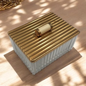Op de afbeelding: Een transparante glazen doos met een gouden deksel en een witte knop. De doos heeft een geribbeld ontwerp en staat op een houten oppervlak.