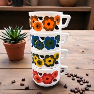 Puede incluir: Una pila de cuatro tazas de café de cerámica blanca con coloridos diseños florales en naranja, azul, amarillo y rojo. Cada taza tiene un asa cuadrada. Las tazas están apiladas sobre una superficie de madera con granos de café esparcidos.