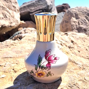 May include: A white ceramic fragrance lamp with a rounded base and a gold-colored metal crown top. The body of the lamp features a hand-painted floral design with a large pink flower, a yellow flower, and green leaves on a smooth, glossy surface.