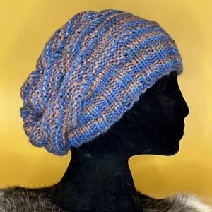 Könnte beinhalten: Eine blaue und braune Strickmütze mit einem lockeren Sitz. Die Mütze sitzt auf einem schwarzen Mannequin-Kopf mit einem grauen Pelzkragen.