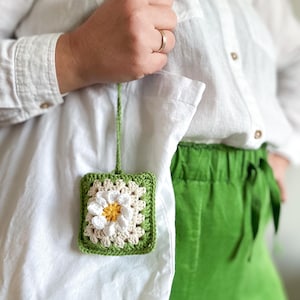 May include: A square, green crocheted charm with a white and yellow daisy design. The charm is attached to a green crocheted cord and hangs on a white tote bag. The person is wearing a white shirt and green pants.