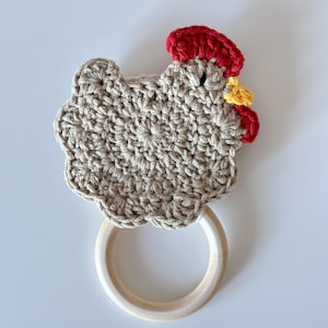 May include: A handmade crochet chicken-shaped teether toy. The chicken is beige with a red comb and wattle, and a yellow beak. It has a wooden ring for easy gripping. The toy is designed for babies and toddlers.