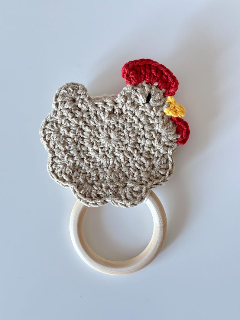 May include: A handmade crochet chicken-shaped teether toy. The chicken is beige with a red comb and wattle, and a yellow beak. It has a wooden ring for easy gripping. The toy is designed for babies and toddlers.