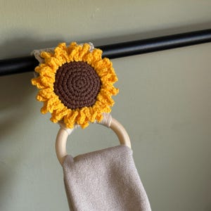 Peut inclure: Un porte-serviettes en forme de tournesol crocheté à la main, avec un anneau en bois et une serviette beige. Le tournesol a un cercle extérieur jaune et un centre marron. Un accessoire charmant pour la cuisine ou la salle de bain.