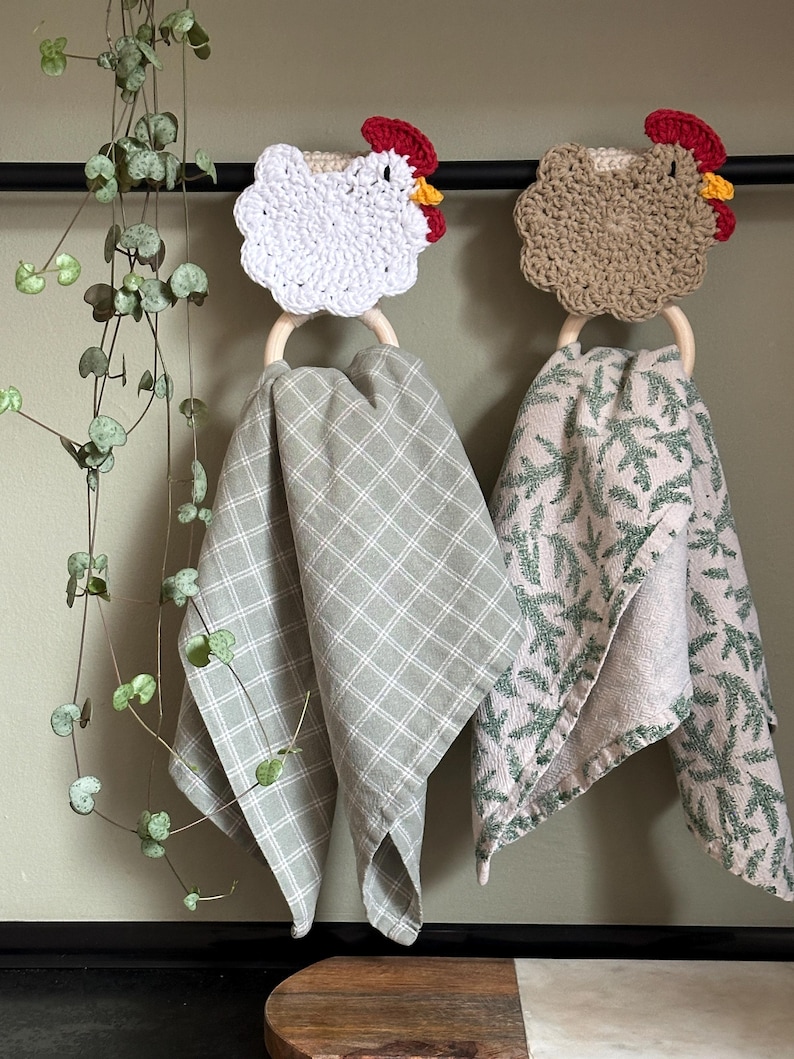 May include: Two crochet chicken-shaped towel holders with wooden rings, each holding a patterned kitchen towel. One chicken is white, the other is tan, both with red combs and yellow wattles. The towels are light green with white plaid and green leaf patterns.