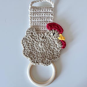 May include: A handmade crochet kitchen towel holder in the shape of a hen. The holder features a beige body, a red comb, and a yellow wattle. It has a wooden ring for hanging and a loop at the top. The towel holder is designed to add a touch of rustic charm to any kitchen.