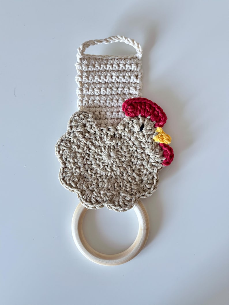 May include: A handmade crochet kitchen towel holder in the shape of a hen. The holder features a beige body, a red comb, and a yellow wattle. It has a wooden ring for hanging and a loop at the top. The towel holder is designed to add a touch of rustic charm to any kitchen.