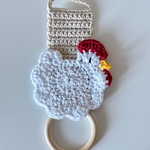 May include: A handmade crochet kitchen towel holder featuring a white chicken design with red and yellow accents. The holder includes a beige loop for hanging and a wooden ring for the towel. The crochet work is detailed and textured.