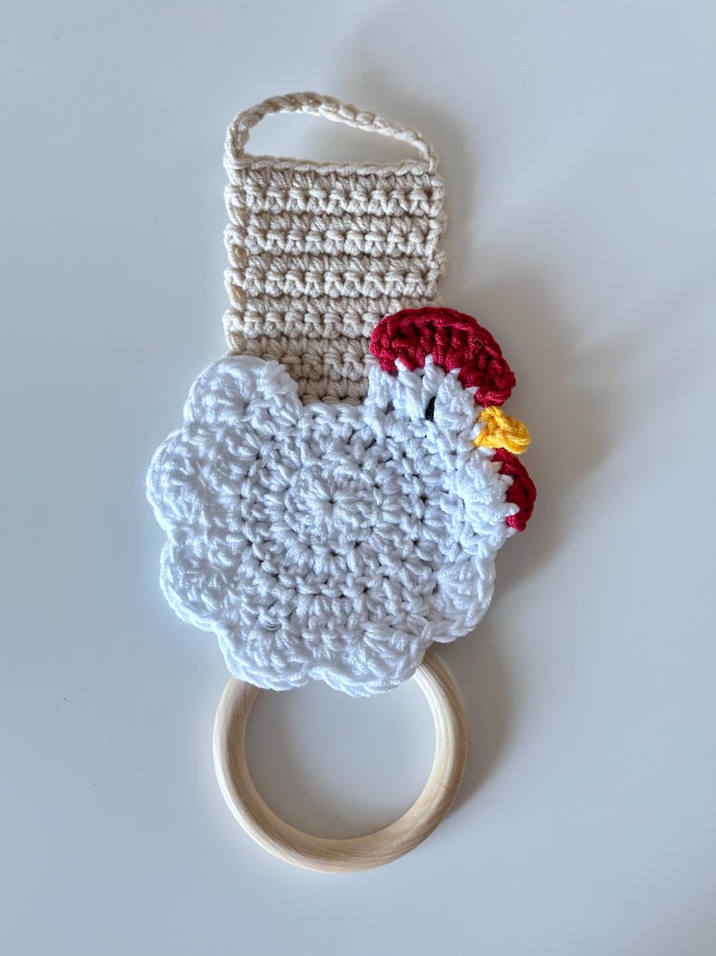 May include: A handmade crochet kitchen towel holder featuring a white chicken design with red and yellow accents. The holder includes a beige loop for hanging and a wooden ring for the towel. The crochet work is detailed and textured.
