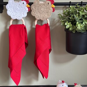 May include: Two crochet chicken towel holders with red napkins hanging below. The chicken heads are white and tan with red combs and yellow beaks. The napkins are a vibrant red color. A black rod holds the holders, with a potted plant to the right.