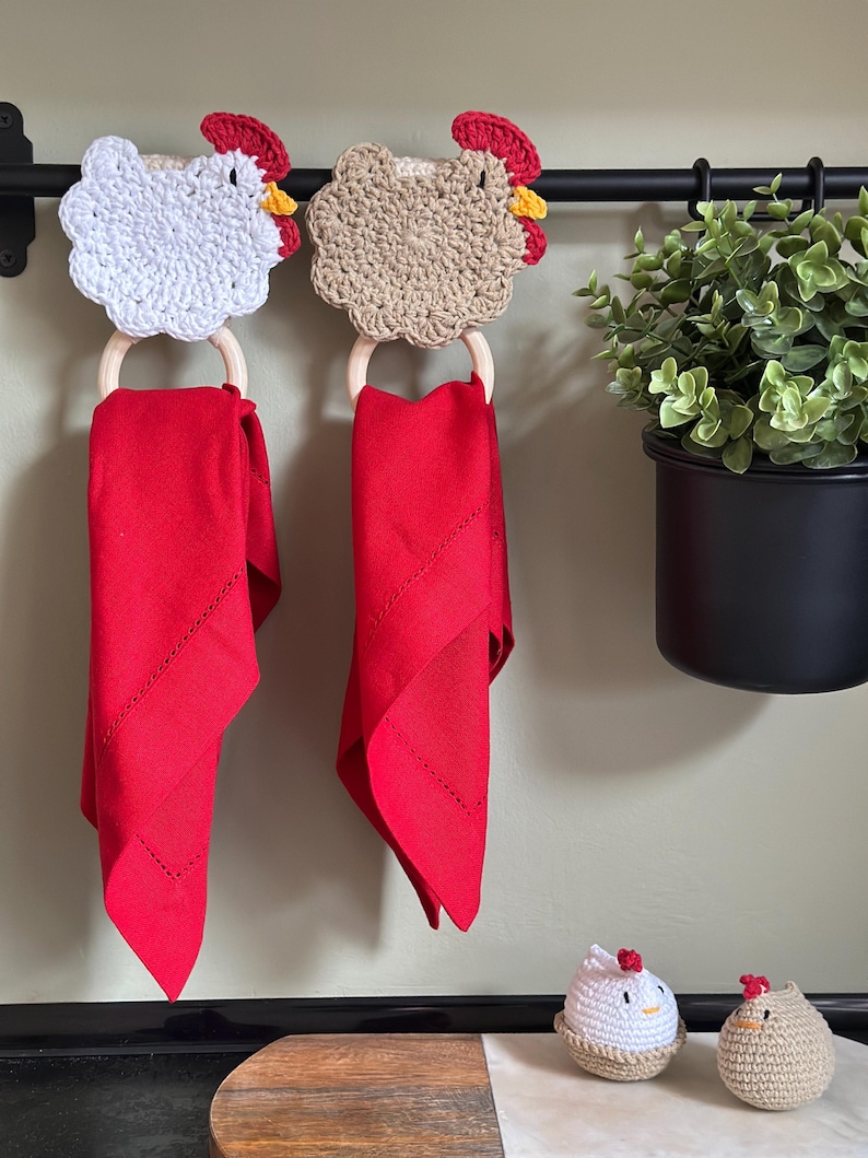 May include: Two crochet chicken towel holders with red napkins hanging below. The chicken heads are white and tan with red combs and yellow beaks. The napkins are a vibrant red color. A black rod holds the holders, with a potted plant to the right.