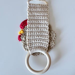 May include: A beige crochet kitchen towel holder with a wooden ring and a loop for hanging. The holder features a red, yellow, and beige crochet chicken design. The towel holder is designed to hold a kitchen towel.