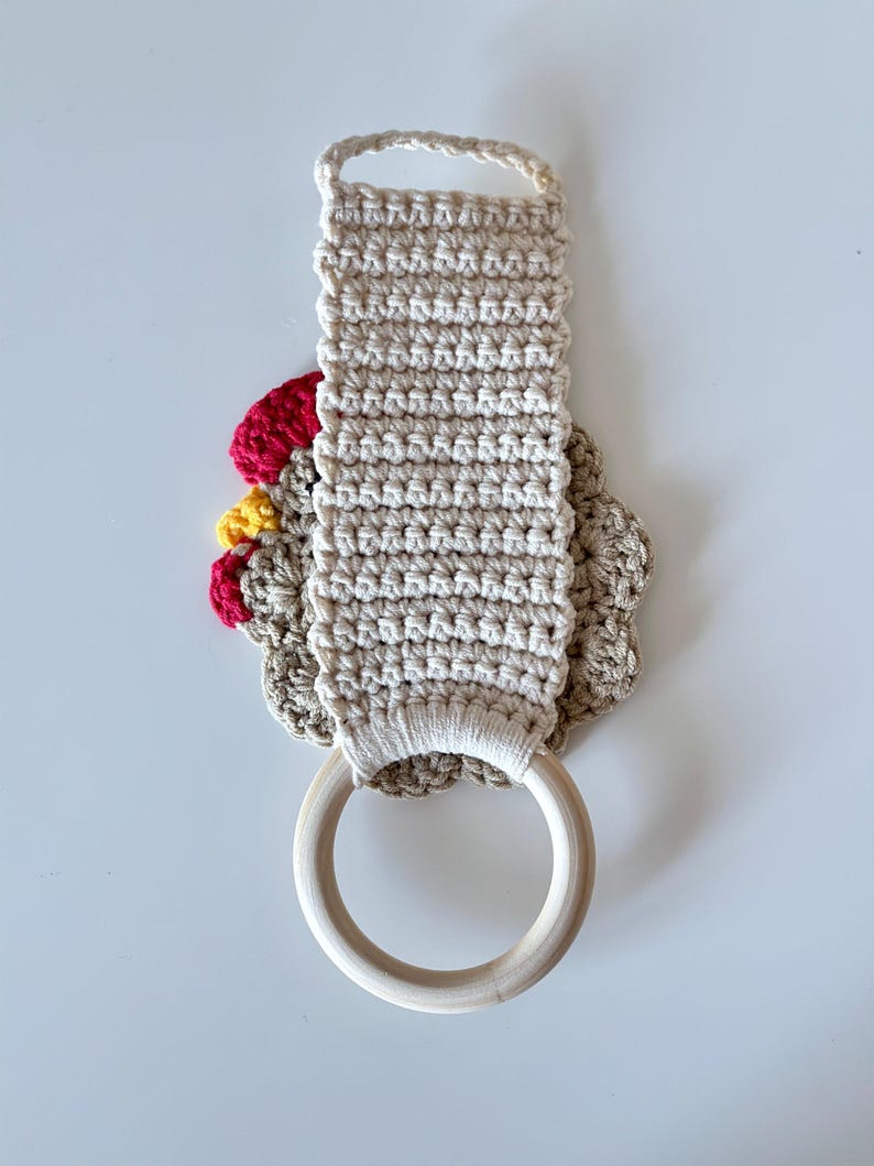 May include: A beige crochet kitchen towel holder with a wooden ring and a loop for hanging. The holder features a red, yellow, and beige crochet chicken design. The towel holder is designed to hold a kitchen towel.