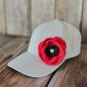 May include: A light gray baseball cap with a vibrant red crocheted poppy flower attached to the front. The poppy has a black and yellow center. The cap is made of a textured fabric.