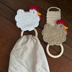 May include: Two handmade crochet kitchen towel holders. One is white, the other is tan, both shaped like chickens with red combs and yellow beaks. Each chicken is attached to a wooden ring. A gray linen towel hangs from one ring.