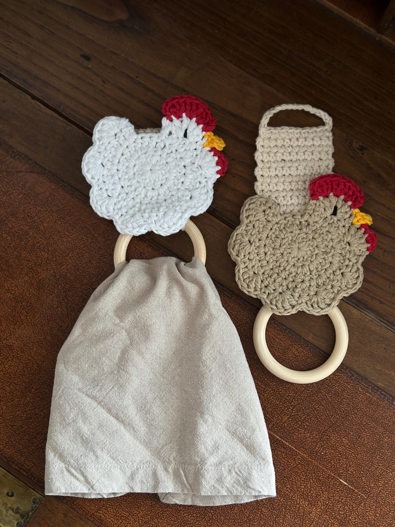 May include: Two handmade crochet kitchen towel holders. One is white, the other is tan, both shaped like chickens with red combs and yellow beaks. Each chicken is attached to a wooden ring. A gray linen towel hangs from one ring.