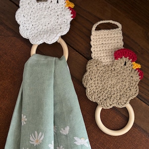 May include: Two crochet chicken-shaped towel holders with wooden rings. One is white, the other is tan. Both have red combs and yellow beaks. A green towel with white floral designs hangs from one ring.