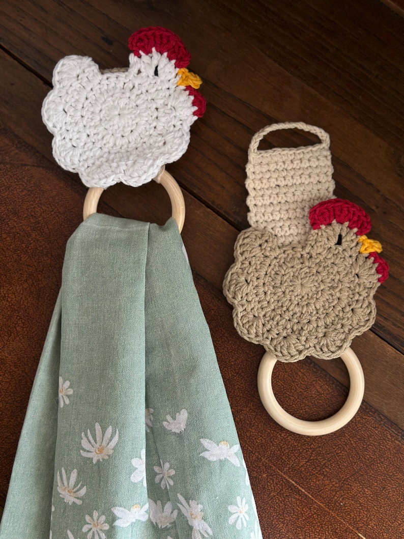 May include: Two crochet chicken-shaped towel holders with wooden rings. One is white, the other is tan. Both have red combs and yellow beaks. A green towel with white floral designs hangs from one ring.