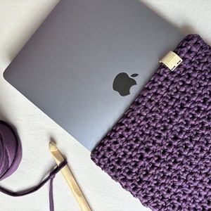 Könnte beinhalten: Eine dunkelviolette, gehäkelte Laptoptasche neben einem silbernen Laptop. Ein Knäuel passender Wolle und eine Häkelnadel aus Holz sind ebenfalls zu sehen. Die Tasche hat ein kleines, rechteckiges Etikett mit der Aufschrift "yarne".