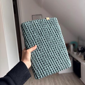 Può includere: Una custodia rettangolare per laptop all'uncinetto verde acqua. La custodia ha un aspetto strutturato e intrecciato e una piccola etichetta color oro sul bordo superiore. La custodia è tenuta da una persona.