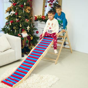 May include: A wooden climbing frame with a red and blue slide. Two children are playing on the slide. The slide is made of wooden slats painted red and blue. The frame is made of natural wood. A Christmas tree is in the background.