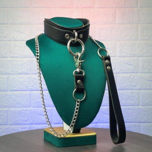 Front angled view of black genuine leather choker and leash set with silver metal hardware displayed on a green mannequin, showing the front O ring connection, chain leash attachment, and leather handle hanging to the side.