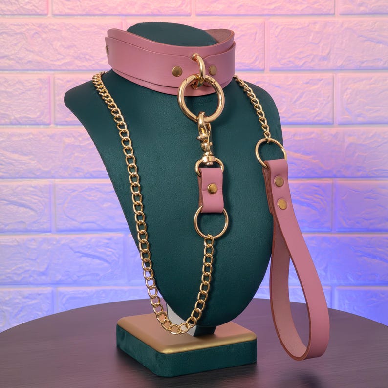 Front angled view of pink genuine leather choker and leash set with gold hardware displayed on a green mannequin, showing the front O ring connection, chain leash attachment, and leather handle hanging to the side.