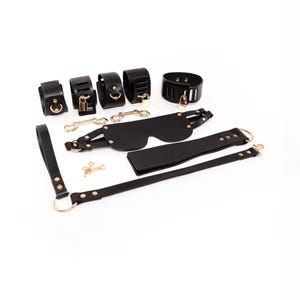 May include: A set of black accessories featuring five cuffs with gold-toned hardware, including two padlocks and keys. The collection includes a blindfold with adjustable straps, a flat paddle, a long strap with a large ring, and two double-ended clips.