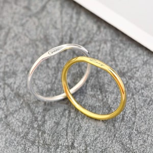 May include: Two thin, smooth metal rings, one silver-toned and one gold-toned, each featuring a delicate, engraved script name on the outer surface. The rings are positioned overlapping each other, showcasing a simple, rounded band design.