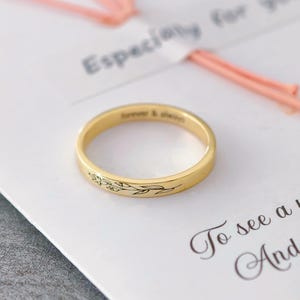 以下が含まれることがあります： 花柄のデザインと「forever & always」の刻印が入ったゴールドリング。リングは「Especially for you」などの文字が書かれた白いカードの上にあります。背景にはピンク色の装飾要素があります。