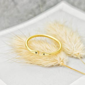 May include: A gold-toned open cuff ring featuring a smooth, polished finish. The two ends of the ring are slightly flattened and each is engraved with a single black letter, specifically the letter M on one side and the letter A on the other.