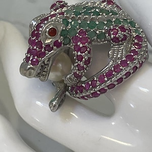 May include: A silver-toned ring shaped like a crocodile, featuring a body encrusted with numerous small, vibrant pink rubies and green emeralds. The crocodile has a single red stone for an eye and holds a smooth, round white pearl in its mouth.