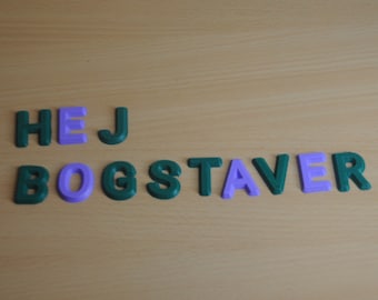 May include: Green and purple plastic letters spelling out the phrase "HEJ BOGSTAVER" on a light brown wooden surface.